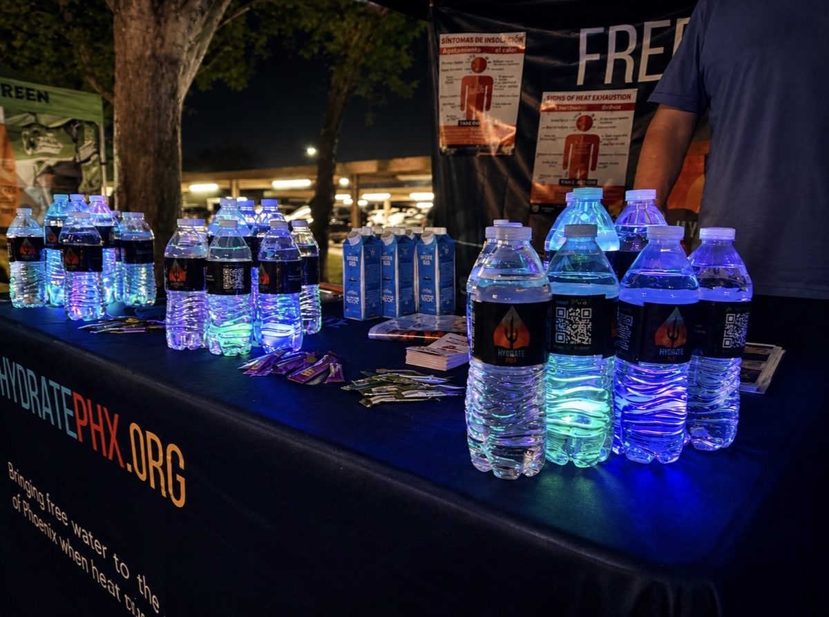 The Hydrate PHX distribution booth at dusk, water bottles and cartons lit up beneath bilingual heat-safety signage.