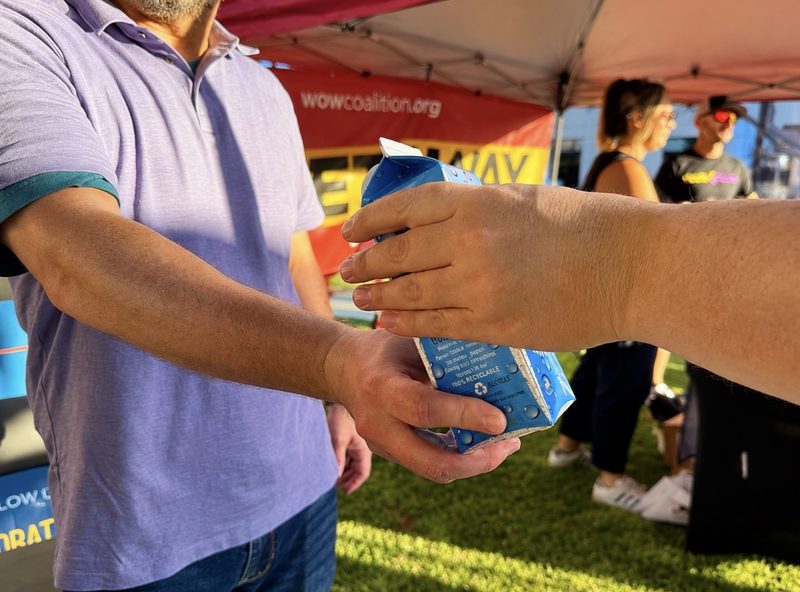 A hand passes a sponsor-branded paper water carton to another hand at a distribution event.